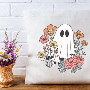 Pu&ograve; includere: Un cuscino bianco con un fantasma e fiori stampati su di esso. Il fantasma &egrave; bianco con due occhi neri ed &egrave; circondato da fiori rosa, arancioni e gialli.