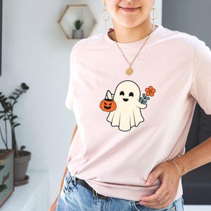 Pu&ograve; includere: Una maglietta rosa con un fantasma bianco che tiene una zucca e un fiore. Il fantasma ha un'espressione felice.
