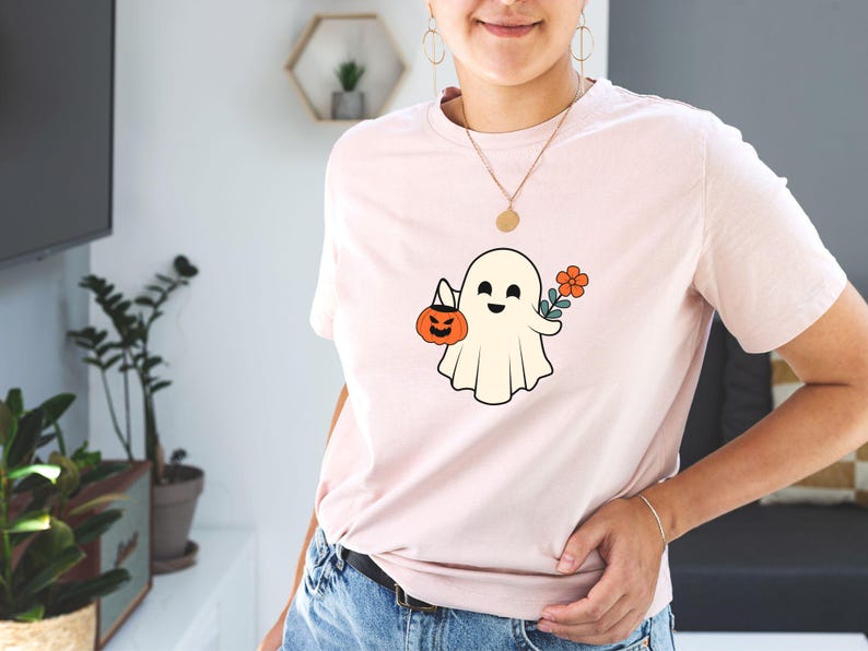 Pu&ograve; includere: Una maglietta rosa con un fantasma bianco che tiene una zucca e un fiore. Il fantasma ha un'espressione felice.