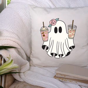 Pu&ograve; includere: Un cuscino bianco con una simpatica illustrazione di un fantasma che tiene due bevande al caff&egrave; freddo rosa e bianco. Il fantasma ha un fiore rosa sulla testa. Il cuscino &egrave; su un divano bianco con una pila di libri.