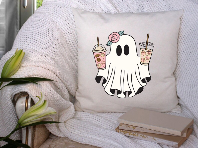 Pu&ograve; includere: Un cuscino bianco con una simpatica illustrazione di un fantasma che tiene due bevande al caff&egrave; freddo rosa e bianco. Il fantasma ha un fiore rosa sulla testa. Il cuscino &egrave; su un divano bianco con una pila di libri.