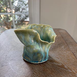 May include: A ceramic vessel with a flared, wavy rim and a cylindrical base. The surface features a reactive glaze with blended tones of sage green, teal, and deep blue, creating a fluid, dripping effect that cascades down the exterior walls.