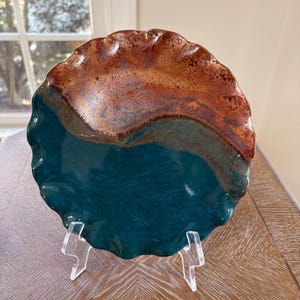 May include: A round ceramic plate with a wavy, scalloped edge. The surface features a two-tone glaze, with a deep teal color covering the bottom half and a textured, mottled rust-brown glaze on the top half, displayed on a clear stand.