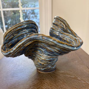 May include: A ceramic bowl with a wavy, organic rim and a textured, rippled surface. The piece features a glaze with swirling patterns of deep blue, light blue, and earthy brown tones, creating a dynamic, fluid appearance throughout.
