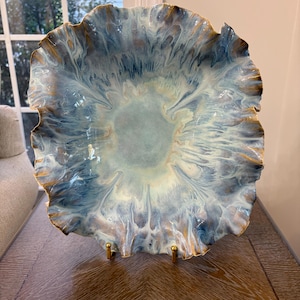 May include: A decorative ceramic bowl with a ruffled, wavy edge. The surface features a marbled glaze in shades of light blue, white, and deep navy, with metallic gold accents along the scalloped rim and throughout the swirling pattern.
