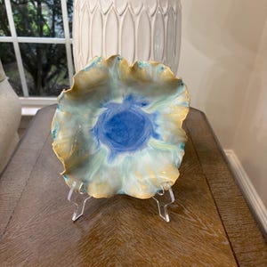 May include: A ceramic bowl with a ruffled, wavy edge featuring a blend of colors including a deep blue center that transitions into soft light blue, cream, and golden yellow tones. The bowl is displayed on a clear acrylic stand.