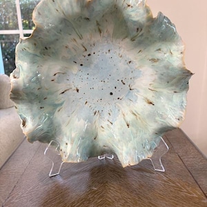 May include: A ceramic bowl with a ruffled, wavy edge featuring a light blue and white glaze. The interior surface is decorated with scattered dark brown speckles and radial streaks that extend from the center toward the scalloped rim.