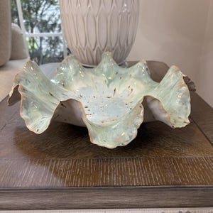 May include: A ceramic bowl with a ruffled, wavy edge featuring a light blue and white glaze with brown speckles. The bowl has a wide, shallow basin and a textured, organic shape that mimics the appearance of a sea shell or a flower petal.