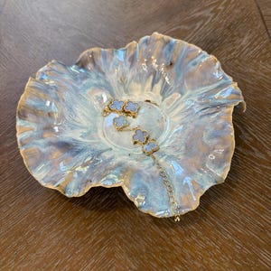 May include: A gold-toned chain bracelet featuring five light blue clover-shaped charms with gold borders, resting inside a ruffled, hand-formed ceramic dish with a glossy, swirling glaze of pale blue, cream, and soft brown tones.