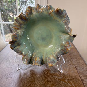 May include: A ceramic bowl with a ruffled, wavy edge featuring a glossy glaze in shades of sage green, light blue, and warm brown accents. The bowl has a smooth, concave center and is displayed on a clear acrylic stand, showcasing its organic form.