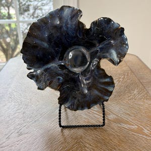 May include: A dark blue and black ceramic sculpture with a ruffled, flower-like shape and a smooth, glossy finish. The piece features a central circular indentation and is displayed on a black metal wire stand that supports its upright position.