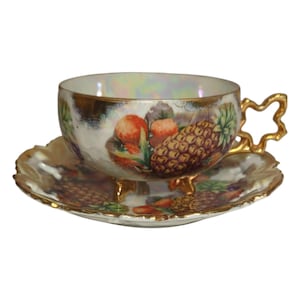 May include: A white porcelain teacup and saucer set with a gold rim and a pineapple and strawberry design. The cup has three gold feet and the saucer has a scalloped edge.