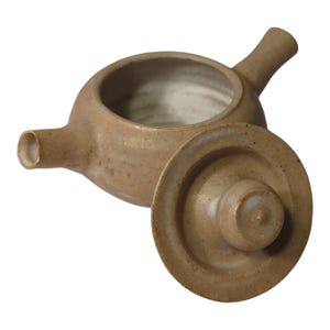 May include: A handmade, tan-colored ceramic teapot with a matching lid. The teapot features a spout, a handle, and a wide opening. The lid is slightly ajar, revealing the interior. The pottery has a rustic, earthy aesthetic.