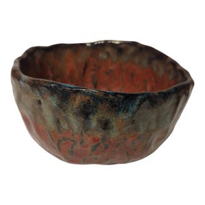 May include: A handmade ceramic bowl with a textured surface. The bowl features a blend of red, brown, and blue-green colors. The rim has an irregular shape, adding to its unique, organic design. This decorative bowl is suitable for serving or display.