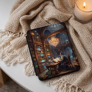 May include: A black tablet case with a detailed illustration of a wizard in a library. The artwork shows a grand library with bookshelves, a chandelier, and the initials "DS" on the desk. The case is resting on a beige knitted blanket.