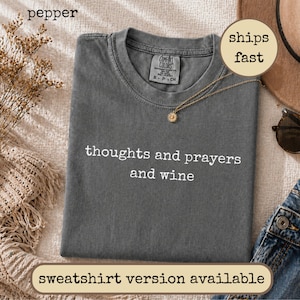 以下が含まれることがあります： 前面に白い文字で thoughts and prayers and wine とプリントされたチャコールグレーの半袖Tシャツ。クルーネックで、襟の内側にComfort Colorsのブランドラベルが付いており、平置きの状態で撮影されています。