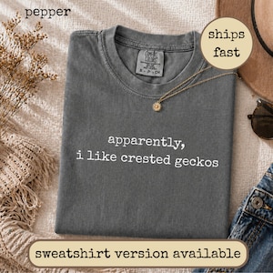 Könnte beinhalten: Ein pfefferfarbenes Kurzarm-T-Shirt mit Rundhalsausschnitt. Das Kleidungsstück ist auf der Brust mit dem weißen Text "apparently, i like crested geckos" bedruckt. Im Kragenbereich befindet sich ein Markenetikett von Comfort Colors.