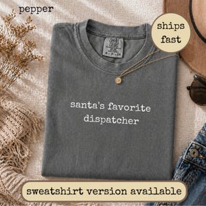 May include: A charcoal grey short-sleeved t-shirt with a crew neckline featuring the white embroidered text 'santa's favourite dispatcher' centred on the chest. The garment has a Comfort Colors brand label inside the collar and a soft texture.