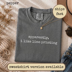 May include: A charcoal gray short-sleeve t-shirt with a crew neckline and a Comfort Colors brand label. The shirt features white text printed on the front that reads, apparently, i like lino printing, in a simple, lowercase typewriter font.