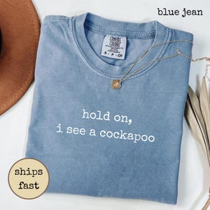 Könnte beinhalten: Ein anthrazitfarbenes Kurzarm-T-Shirt mit dem weißen Text "hold on, i see a cockapoo" auf der Vorderseite. Das Kleidungsstück hat einen Rundhalsausschnitt und ein Comfort Colors Markenetikett am Innenkragen und zeigt ein schlichtes Design.