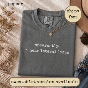 May include: A charcoal gray short-sleeve t-shirt with the white text "apparently, i hear lateral lisps" printed on the front. The shirt features a Comfort Colors brand label at the inner neckline and a crew neck collar design.
