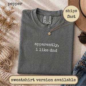 May include: A charcoal grey short-sleeve t-shirt with the white embroidered text "apparently, i like dnd" centred on the chest. The shirt features a crew neckline and a Comfort Colors brand label on the inside collar.