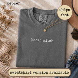 Könnte beinhalten: Ein anthrazitfarbenes Kurzarm-T-Shirt mit Rundhalsausschnitt und dem in weißem Garn auf die Brust gestickten Text basic witch. Das T-Shirt verfügt über ein Comfort Colors Markenetikett am Innenkragen und eine weiche, verwaschene Stoffstruktur.