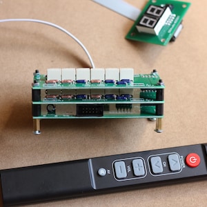 Könnte beinhalten: Eine grüne Platine mit mehreren Knöpfen und einem digitalen Display. Die Platine ist mit einer schwarzen Fernbedienung mit Knöpfen für Strom, Kanal, Lautstärke und AV verbunden.