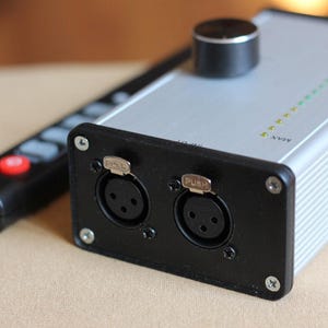 Könnte beinhalten: Silbernes Audiogerät mit schwarzer Frontplatte mit zwei XLR-Anschlüssen und einem Lautstärkeregler. Eine schwarze Fernbedienung mit roter Taste befindet sich im Hintergrund. Das Gerät hat eine Reihe grüner und gelber Kontrollleuchten.