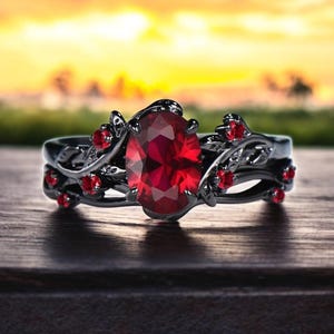 Può includere: Un anello nero con una grande pietra ovale rossa circondata da pietre rosse più piccole e un design a vite.