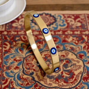 May include: A gold-colored bangle bracelet with four blue and white evil eye charms. The bracelet is smooth and reflective, set against a patterned red, blue, and beige rug. The bracelet is a piece of jewelry.