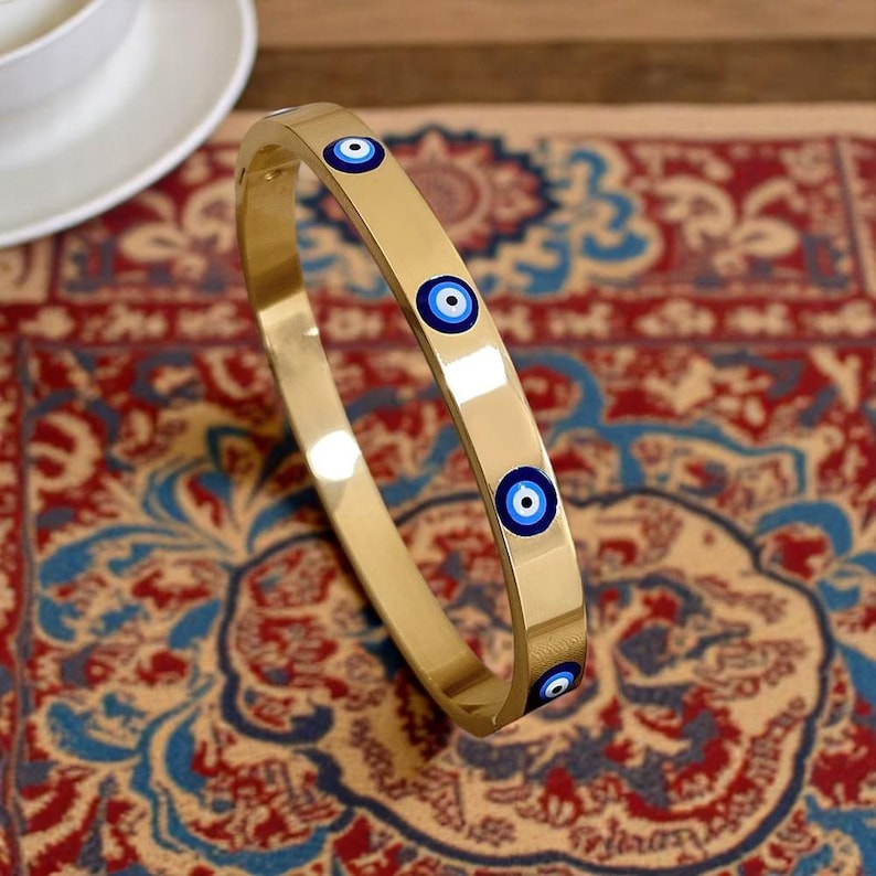 May include: A gold-colored bangle bracelet with four blue and white evil eye charms. The bracelet is smooth and reflective, set against a patterned red, blue, and beige rug. The bracelet is a piece of jewelry.