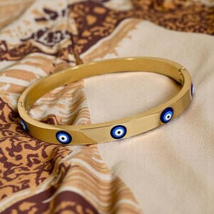May include: A gold-colored bangle bracelet with multiple blue and white evil eye charms. The bracelet is made of a shiny, smooth metal and is resting on a patterned fabric.