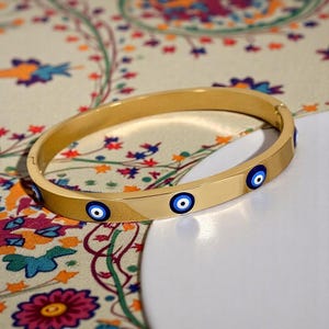 May include: A gold-colored bangle bracelet with multiple blue and white evil eye charms. The bracelet is oval-shaped and has a clasp closure. The background features a patterned fabric with floral and abstract designs.