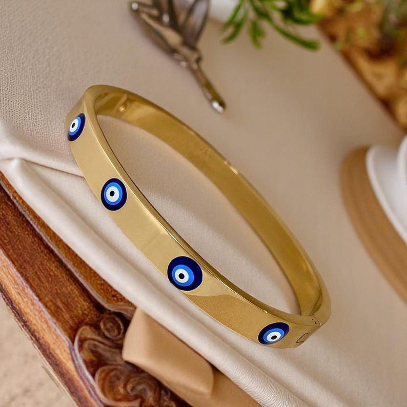 May include: Gold-toned bangle bracelet with blue and white evil eye charms. The bracelet has a smooth, polished surface and a simple, elegant design. The evil eye charms are evenly spaced around the bracelet.
