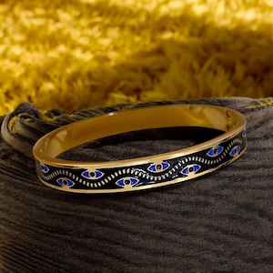 May include: A gold bangle bracelet with a black enamel band and a repeating pattern of blue evil eyes.