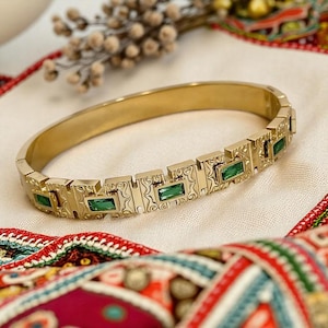 Puede incluir: Un brazalete de oro con un diseño decorativo y piedras preciosas verdes. El brazalete está sobre un fondo de tela estampada.