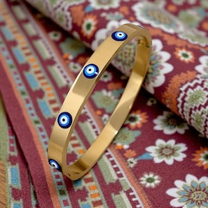 May include: Gold-toned bangle bracelet featuring multiple blue and white evil eye charms. The bracelet has a smooth, polished finish and is displayed on a patterned fabric with red, green, and orange floral designs.