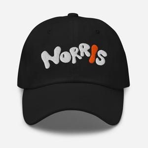 Pode incluir: Boné de beisebol preto com o nome "Norris" bordado em branco, com a letra "i" em laranja. O boné tem uma aba curva e uma alça ajustável.