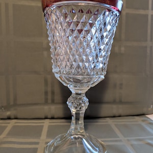 May include: A clear, vintage-style goblet with a diamond-cut pattern on the bowl and a red rim. The stem and base are also clear, creating an elegant and classic look. The goblet is likely made of glass.