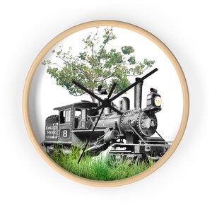 May include: A round wall clock with a light wood frame. The clock face features a black and white illustration of a vintage steam train with a tree growing from the top. Green grass is at the bottom of the image.