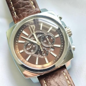 May include: A Thomas Sabo chronograph watch with a square silver-tone stainless steel case, a brown dial, and three sub-dials. The watch features silver-tone hour markers, a date window at the four o'clock position, and a textured brown leather strap.