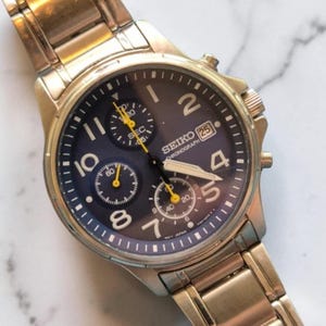 May include: A gold-toned Seiko Chronograph watch with a dark blue face. The watch features multiple sub-dials with yellow accents, white numerals, and a date display at the 3 o'clock position. The watch band is also gold-toned.