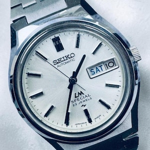 May include: A silver-toned Seiko LM Special automatic wristwatch. The watch face is white with black hands and markers, and a day-date display showing "SAT 10." The watch band is a matching silver metal. The watch has "23 Jewels" and "Japan 5216" printed on the face.