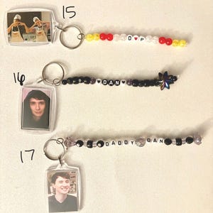 May include: Three rectangular acrylic photo keychains labeled 15, 16, and 17. Keychain 15 features a photo of two people and a beaded strand with red, yellow, and white beads spelling D heart P. Keychain 16 has a photo of a person and black beads spelling DAN. Keychain 17 has a photo of a person and beads spelling DADDY DAN.