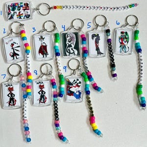 May include: A collection of keychains featuring cartoon characters. Each keychain has a clear rectangular charm with a character image and a beaded lanyard. The lanyards are multi-colored and spell out names like "BLITZO" and "FIZZAROLLI". One keychain has the text "HELLUVA LOT OF PRIDE".
