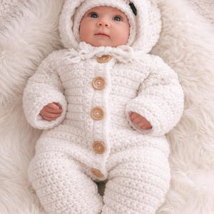 Op de afbeelding: Een baby in een witte, gehaakte berenpak met een capuchon en bijpassende hoed. Het pak heeft houten knopen. De baby ligt op een zacht, wit oppervlak.