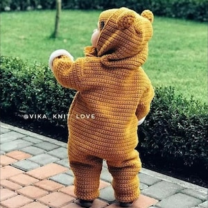 Pode incluir: Um bebê vestindo um macacão de crochê marrom com formato de urso, com capuz e orelhas de urso. O macacão tem uma borda branca ao redor dos pulsos e tornozelos. O bebê está em pé em um caminho de paralelepípedos.