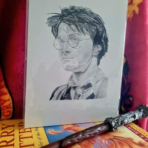 Puede incluir: Un boceto a lápiz de un joven con gafas redondas, con una varita y un libro en el fondo. La varita es marrón con un mango plateado detallado. El libro tiene una cubierta amarilla con el título "Harry Potter".