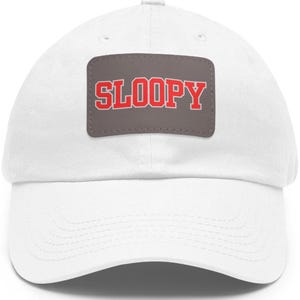 Puede incluir: Gorra de béisbol blanca con visera curva. Un parche rectangular en la parte delantera muestra la palabra "SLOOPY" en rojo, con contorno blanco, sobre un fondo gris. La gorra es de tela suave.